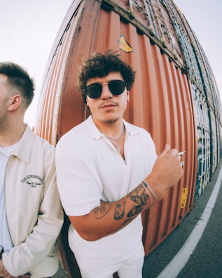 A young man with sunglasses and tattoos on his left arm is standing next to a rust-colored shipping container, wearing a white shirt and holding a cigarette. Another person is partially visible, facing away and wearing a jacket with the words 'The Stronghold' printed on it.
