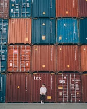 A person stands casually in front of a large stack of colorful shipping containers. The containers are organized in neat rows, featuring hues of red, blue, and orange. Each container has various labels and markings typically seen in industrial settings.