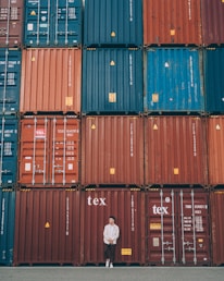 A person stands casually in front of a large stack of colorful shipping containers. The containers are organized in neat rows, featuring hues of red, blue, and orange. Each container has various labels and markings typically seen in industrial settings.