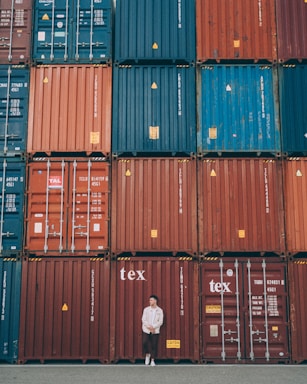 A person stands casually in front of a large stack of colorful shipping containers. The containers are organized in neat rows, featuring hues of red, blue, and orange. Each container has various labels and markings typically seen in industrial settings.