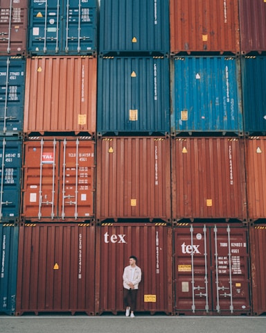 A person stands casually in front of a large stack of colorful shipping containers. The containers are organized in neat rows, featuring hues of red, blue, and orange. Each container has various labels and markings typically seen in industrial settings.