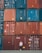A person stands casually in front of a large stack of colorful shipping containers. The containers are organized in neat rows, featuring hues of red, blue, and orange. Each container has various labels and markings typically seen in industrial settings.