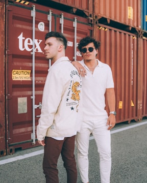 Two men are standing in front of large red shipping containers. One man is wearing a white jacket with a tiger graphic on the back and brown pants, while the other man is dressed in a white polo and pants, accessorized with sunglasses and rings. The scene has an industrial feel with the containers as the backdrop.