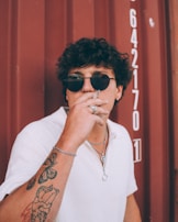 A person with curly hair and wearing sunglasses and a white collared shirt smokes a cigarette. Their arm is adorned with tattoos, and they are accessorized with multiple rings and a necklace. The background features a maroon surface with numbers, suggesting an industrial setting.