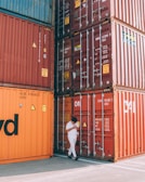 A person dressed in white walks beside stacked shipping containers in various shades of orange and red. The containers are labeled with shipping information and numbers. The scene takes place outdoors, with a concrete ground visible beneath the containers.