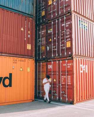 A person dressed in white walks beside stacked shipping containers in various shades of orange and red. The containers are labeled with shipping information and numbers. The scene takes place outdoors, with a concrete ground visible beneath the containers.