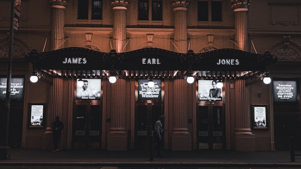 A nighttime view of a theater entrance illuminated by warm lights, featuring prominent signage with the names 'James Earl Jones' above the doors. Below the signs, there are lit posters with images of people and text advertising theatrical performances. The facade is ornate, with pillars and decorative elements, and a few people are seen walking by.