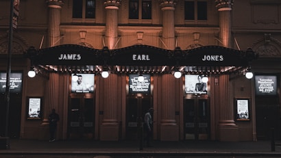 A nighttime view of a theater entrance illuminated by warm lights, featuring prominent signage with the names 'James Earl Jones' above the doors. Below the signs, there are lit posters with images of people and text advertising theatrical performances. The facade is ornate, with pillars and decorative elements, and a few people are seen walking by.
