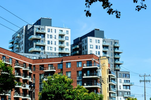 A cluster of modern apartment buildings, featuring a combination of brick and concrete materials. The structures are multi-storied with balconies. The foreground displays greenery and trees, along with power lines crossing horizontally. The sky is clear and blue.