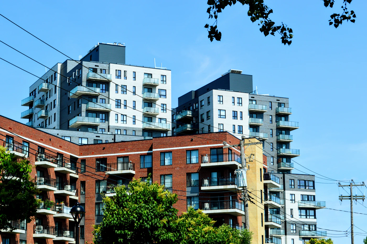 Mid-rise residential apartment buildings with rows of balconies above older red brick walk-ups in a Montreal neighbourhood under a blue sky