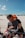 A couple sitting on a red ATV in a sandy desert area. The woman is wearing a bright orange hat and sunglasses, smiling and hugging the man from behind. The man also has sunglasses and is holding the handlebars. The sky is clear with a few fluffy clouds.