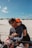 A couple sitting on a red ATV in a sandy desert area. The woman is wearing a bright orange hat and sunglasses, smiling and hugging the man from behind. The man also has sunglasses and is holding the handlebars. The sky is clear with a few fluffy clouds.