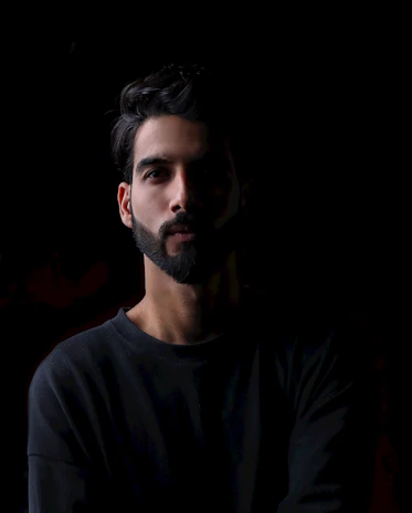 Elegant man with a sharp haircut and beard, illuminated by golden light against a deep black background.