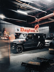 A black SUV is parked inside a garage or repair shop which has a high ceiling with visible metal beams and fluorescent lights. A large sign with the word 'Dayton' is displayed on the back wall. There's another vehicle, white in color, parked nearby. Various tools and boxes are scattered around on the garage floor. A road work sign is reflected on the glass, adding depth to the scene.