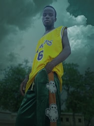 A person stands confidently outdoors, wearing a yellow sports jersey with the number 6 and green pants featuring a cartoon character design. The background consists of a dramatic, cloudy sky and some trees.