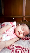 A baby is peacefully sleeping on a patterned pillow, wearing a pink sleeveless top with a cat illustration. The background consists of wooden furniture, giving a warm, cozy setting.