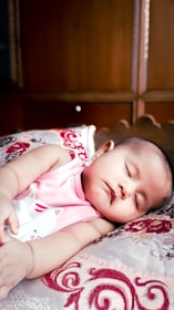 A baby is peacefully sleeping on a patterned pillow, wearing a pink sleeveless top with a cat illustration. The background consists of wooden furniture, giving a warm, cozy setting.