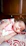 A baby is peacefully sleeping on a patterned pillow, wearing a pink sleeveless top with a cat illustration. The background consists of wooden furniture, giving a warm, cozy setting.