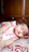 A baby is peacefully sleeping on a patterned pillow, wearing a pink sleeveless top with a cat illustration. The background consists of wooden furniture, giving a warm, cozy setting.