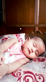 A baby is peacefully sleeping on a patterned pillow, wearing a pink sleeveless top with a cat illustration. The background consists of wooden furniture, giving a warm, cozy setting.