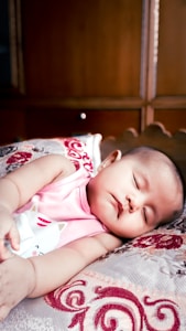 A baby is peacefully sleeping on a patterned pillow, wearing a pink sleeveless top with a cat illustration. The background consists of wooden furniture, giving a warm, cozy setting.