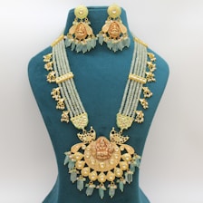 An ornate necklace set displayed on a teal jewelry bust. The necklace features intricate gold designs with multiple strands of pale green beads and elaborate gold ornaments. It is accompanied by matching earrings that share the same gold detailing and dangling beads.