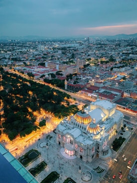 An aerial view of a cityscape features a prominent, ornately designed building with a colorful domed roof, beautifully lit at dusk. The building is surrounded by bustling streets and lush green trees, with city structures extending into the horizon. The sky carries a hint of sunset colors, adding warmth to the scene.