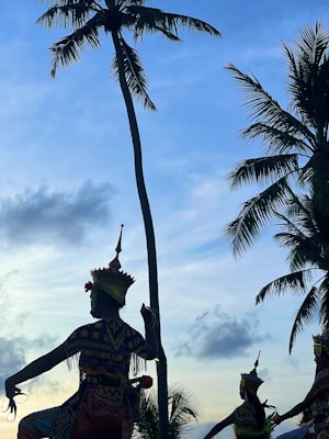 Silhouetted figures wear traditional, elaborate costumes and headdresses, performing a cultural dance against a backdrop of tall palm trees and a sky transitioning from daylight to evening, with scattered clouds.