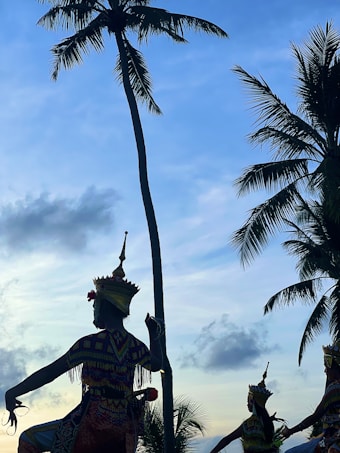 Silhouetted figures wear traditional, elaborate costumes and headdresses, performing a cultural dance against a backdrop of tall palm trees and a sky transitioning from daylight to evening, with scattered clouds.