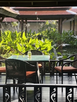 A serene outdoor setting featuring a glass-top table with a single decorative object, surrounded by four wicker chairs with orange cushions. The foreground includes intricate wooden railing designs, while the background is lush with green foliage and traditional architecture with red-tiled roofs.