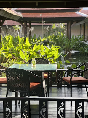 A serene outdoor setting featuring a glass-top table with a single decorative object, surrounded by four wicker chairs with orange cushions. The foreground includes intricate wooden railing designs, while the background is lush with green foliage and traditional architecture with red-tiled roofs.