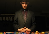 A man in a suit stands over a tabletop filled with colorful game pieces. The lighting is dim, creating a dramatic and contemplative atmosphere. His hands are clasped in front of him, and he appears to be deep in thought.