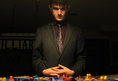 A man in a suit stands over a tabletop filled with colorful game pieces. The lighting is dim, creating a dramatic and contemplative atmosphere. His hands are clasped in front of him, and he appears to be deep in thought.