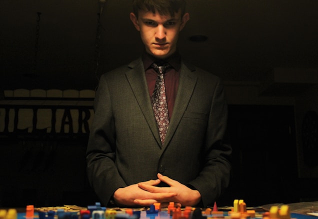 A man in a suit stands over a tabletop filled with colorful game pieces. The lighting is dim, creating a dramatic and contemplative atmosphere. His hands are clasped in front of him, and he appears to be deep in thought.