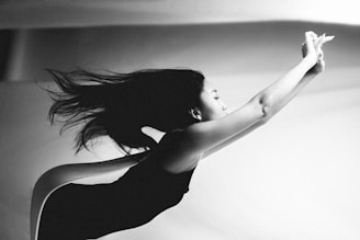A black and white photograph capturing a woman in motion, with her body stretched forward and hair flowing behind. The composition emphasizes a sense of grace and fluidity, as light and shadow highlight her form against a soft, out-of-focus background.