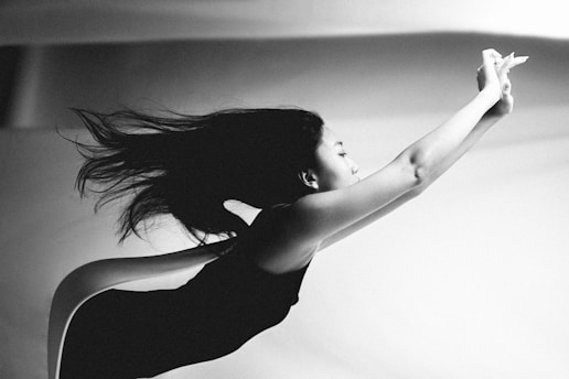A black and white photograph capturing a woman in motion, with her body stretched forward and hair flowing behind. The composition emphasizes a sense of grace and fluidity, as light and shadow highlight her form against a soft, out-of-focus background.