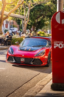 A sleek, red sports car is parked on a sunlit street next to an Australia Post mailbox. The background features people dining outdoors at a café with lush greenery and trees providing shade. The atmosphere is vibrant and relaxed.