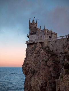 A picturesque castle perched on the edge of a steep cliff overlooking the vast ocean. The sky is a blend of blues and soft oranges, suggesting a sunset or sunrise, casting a serene light on the structure. The castle features gothic architecture with multiple turrets and flags, creating an enchanting and historical ambiance.