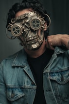 A person wearing a detailed, steampunk-style mask that resembles a metallic skull with gears. The mask features round, goggle-like eye covers with a mechanical design and intricate metal details. The person is dressed in a denim jacket and has a hand resting on their shoulder.