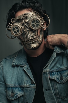 A person wearing a detailed, steampunk-style mask that resembles a metallic skull with gears. The mask features round, goggle-like eye covers with a mechanical design and intricate metal details. The person is dressed in a denim jacket and has a hand resting on their shoulder.