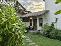 A cozy outdoor patio area surrounded by lush green plants and trees. The building has a rustic appearance with a thatched roof and wooden doors. A small chair and some pots are placed near the entrance, and a stone path leads up to the door. The sky is partly cloudy, adding a serene backdrop to the scene.