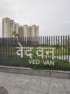 A tall set of residential buildings stand behind a fence with the words 'Ved Van' written on it. The sky above is overcast, and the scene features green foliage and well-maintained lawns in the foreground.