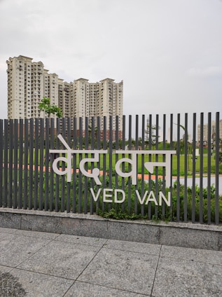 A tall set of residential buildings stand behind a fence with the words 'Ved Van' written on it. The sky above is overcast, and the scene features green foliage and well-maintained lawns in the foreground.
