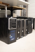 Several desktop computer towers are lined up on the floor next to each other. The computers are placed under a table in an indoor setting. The visible brands include Acer and other unspecified brands. The towers have multiple drive slots and ports visible on the front panels.