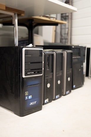 Several desktop computer towers are lined up on the floor next to each other. The computers are placed under a table in an indoor setting. The visible brands include Acer and other unspecified brands. The towers have multiple drive slots and ports visible on the front panels.