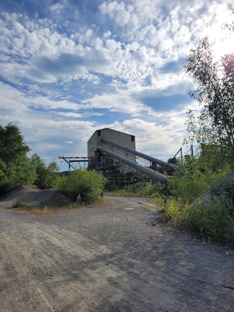 An industrial structure stands amidst lush green vegetation under a sky filled with scattered clouds. The building features a slanted conveyor belt extending from its side, suggesting a quarry or processing facility. The foreground includes a dirt road leading towards the structure.