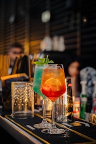 Two colorful cocktails are placed prominently on a bar counter with a blurred background indicating a bar setting. One drink is in a clear glass with an orange hue, garnished with a lemon slice and mint leaves, while the other has a blue-green color.