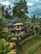 A two-story house made of natural materials, possibly bamboo, is nestled in a lush tropical setting. Tall palm trees and dense greenery surround the house, creating a serene and secluded atmosphere. A hammock hangs on the lower level porch, and stone steps lead up to the entrance where a person stands, dressed casually.