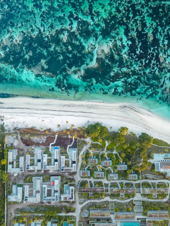 An aerial view of a coastline with turquoise waters meeting a sandy beach. The shoreline is lined with modern buildings, likely part of a resort, surrounded by greenery and pathways. The ocean is vibrant with dark and light blue hues, creating an inviting and serene landscape.