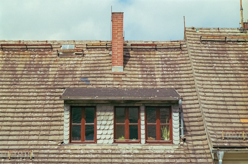 An old, weathered roof with overlapping shingles that have signs of wear and moss growth. A small brick chimney protrudes from the roof. There is a dormer window with two large windows, accented by white framing. Some green plants are visible on the windowsill.